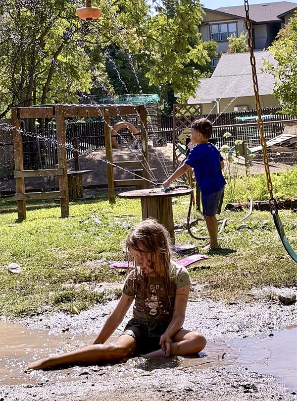 Two young children play outside on the grass and the mud. A hose and sprinkler shoot water into the air.
