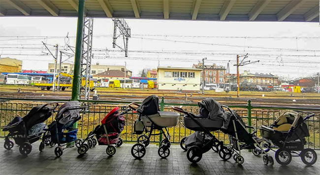 Strollers on a train platform. Train tracks and low buildings are visible in the distance.