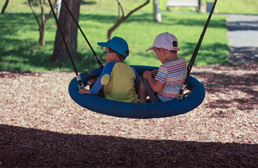 Two preschool-aged boys in brightly-colored t-shirts and baseball hats sit in a tire-style tree swing on a sunny day.