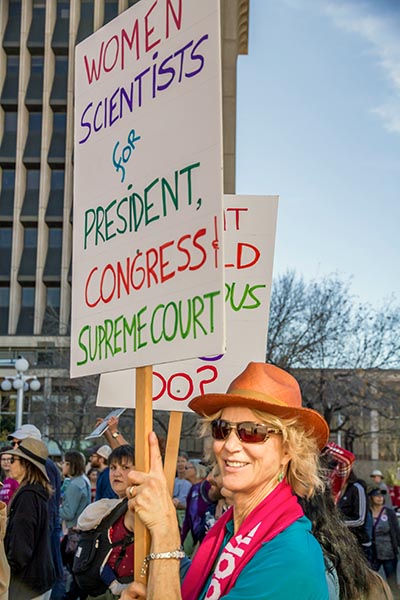 Middle-aged woman holding up a protest sign saying, "Women Scientist for President, Congress, Supreme Court"