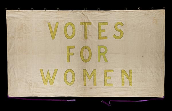 An antique cloth banner with Votes For Women sewn on in yellow capital letters