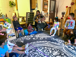 A teacher and preschool-aged children discuss the 6 persona dolls the teacher is holding as they all sit around a large area rug in their classroom