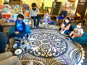 Preschool-aged children sit on an area rug in their classroom talking about Evan, the persona doll who is in a toy-sized wheelchair