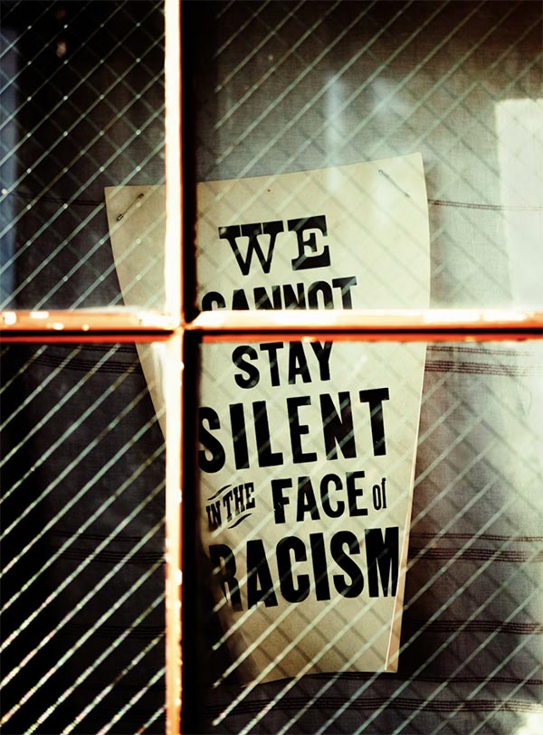 A sign reading, "We cannot stay silent in the face of racism," is seem behind a wire-reinforced window.