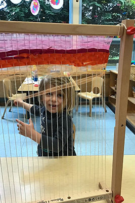 Pre-school girl seen through the strings of a table-top loom as she weaves