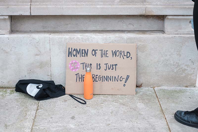Handwritten sign on cardboard leaning against a cement step reading Women of the world, this is just the beginning. Next to the sign on the ground is a black canvas bag and an orange metal water bottle