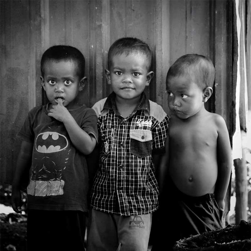 Black-and-white image of 3 black toddler boys. One is shirtless, one wears a batman shirt, and the boy in the middle wears a plaid shirt.