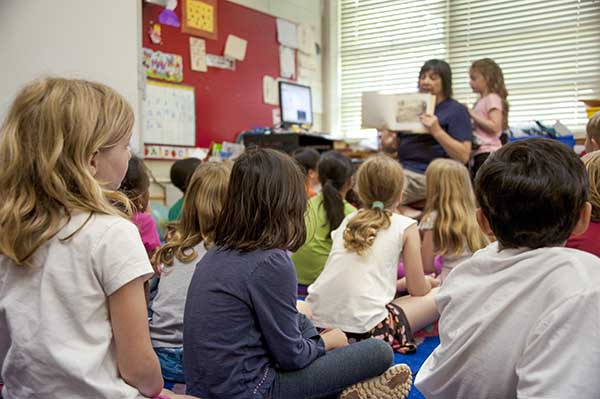 A teacher holds up a book for a group of early elementary-aged students seated on the ground in front of her in the classroom.