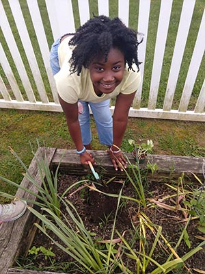 A young girl looks up and smiles. She is holding a small garden shovel and digs in the dirt of a raised garden bed.